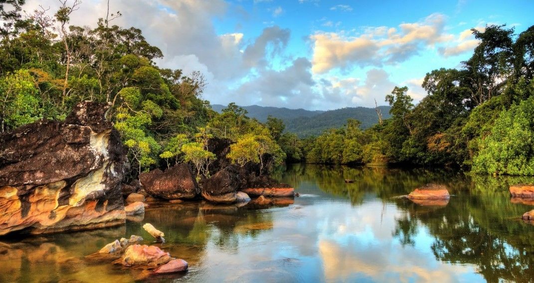 Scenic view of a tranquil river with large rocks and lush greenery under a cloudy blue sky.